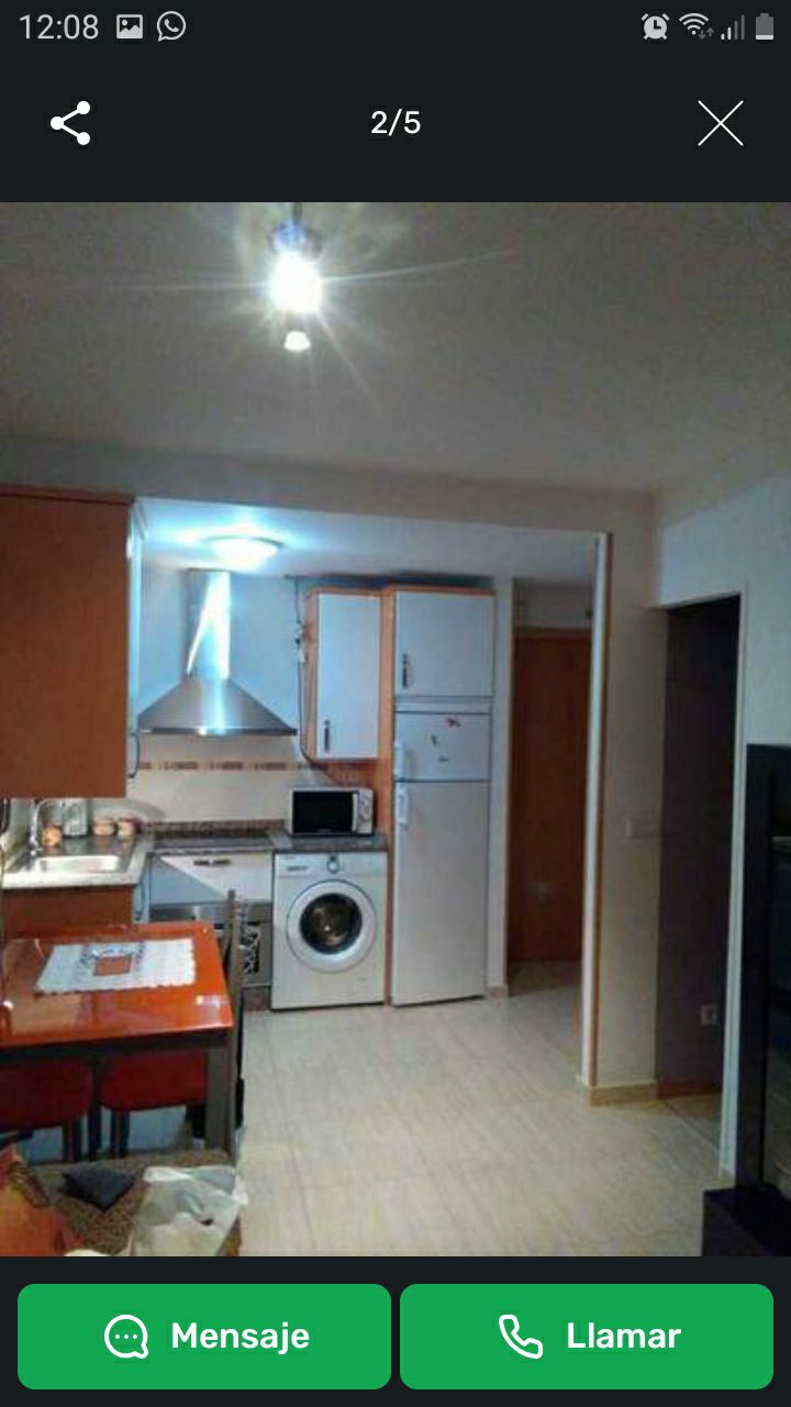 A compact kitchen area is visible, featuring essential appliances such as a refrigerator, washing machine, and stovetop. A dining table with orange accents is placed nearby. The space is illuminated by a ceiling light, enhancing the overall brightness of the kitchen.