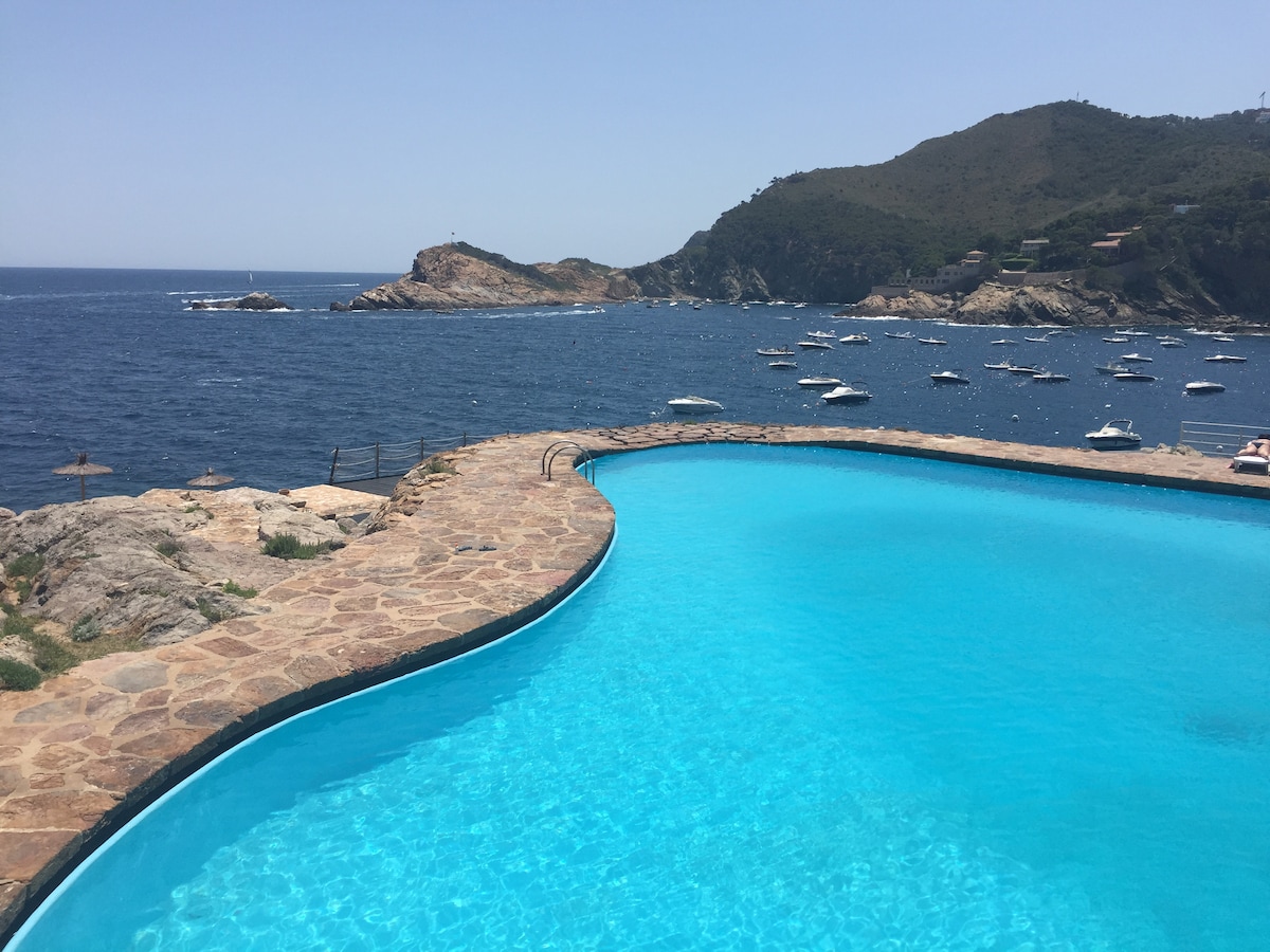 A large salt-water pool is featured, with its turquoise water contrasting against the rugged coastline and the blue sea. Rocky outcroppings and boats are visible in the background, highlighting the scenic marine surroundings.