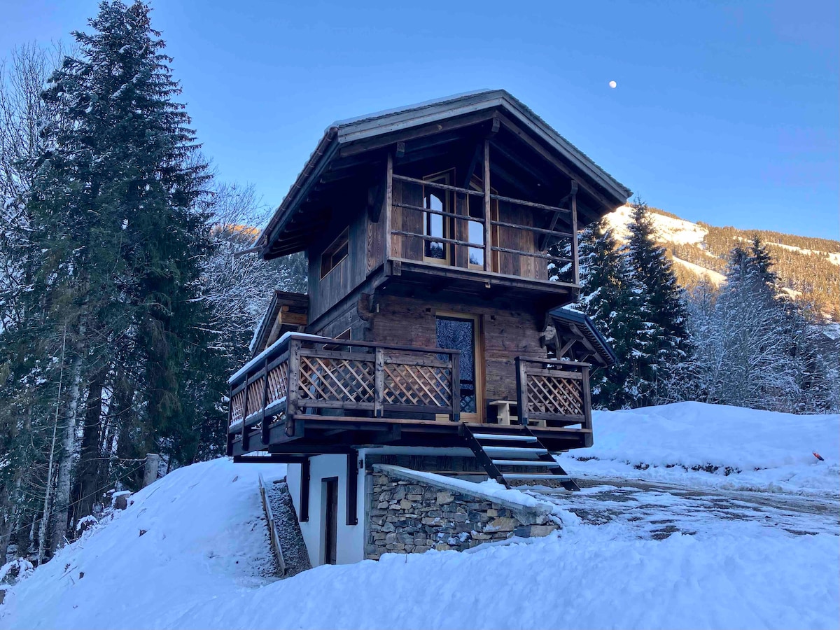 A traditional chalet is set against a snowy landscape, featuring a wooden exterior and two balconies. The structure is elevated, with stairs leading to the entrance. Tall evergreen trees surround the property, and the sky displays a hint of blue with a visible moon.
