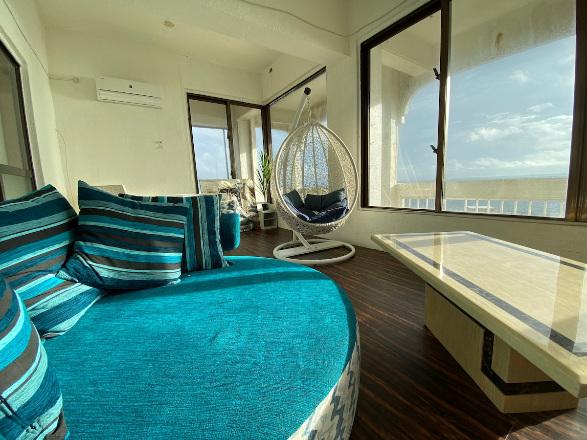 A spacious living area features a circular blue seating arrangement with striped cushions. A suspended chair adds a relaxed touch, while large windows allow natural light and views of the sky. A light-colored table complements the modern decor.