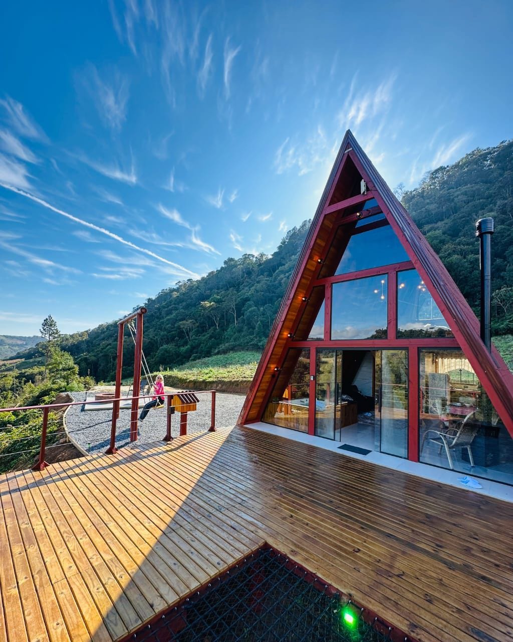 A unique A-frame structure features large windows, allowing for ample natural light. The outdoor deck is constructed of wooden planks and includes a seating area. Lush greenery surrounds the property, enhancing the serene environment. A clear blue sky adds to the inviting setting.