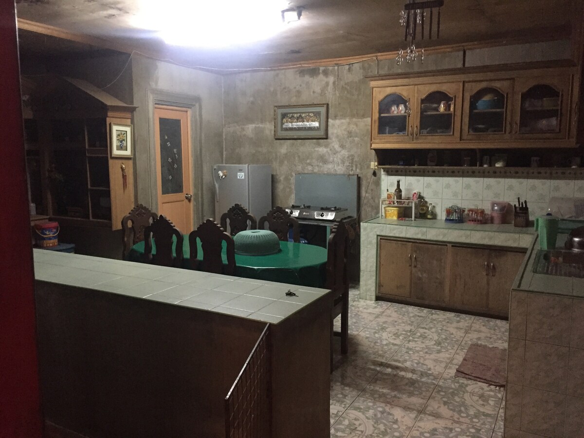 A spacious kitchen area is equipped with wooden cabinetry and a large countertop. Two dining tables surround a central seating arrangement. The room is illuminated by ceiling lights, creating a functional space for meals. Appliances include a refrigerator and stove, with tiled flooring enhancing the layout.