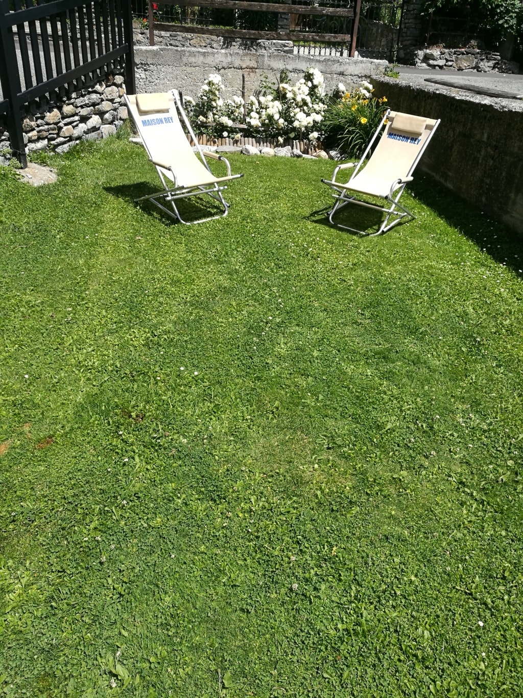 A grassy outdoor area features two reclining chairs positioned for relaxation. A flowerbed with a variety of blooms adds color to the setting, surrounded by stone borders. The space provides a tranquil environment for enjoying fresh air.