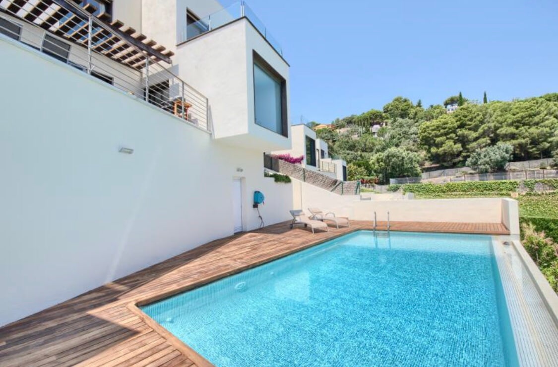 A private pool is framed by a wooden deck, surrounded by lush greenery. The clear water reflects the blue sky above, while sun loungers are positioned nearby. The building’s modern architecture is visible in the background, highlighting large windows that showcase the scenic environment.
