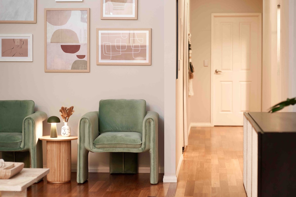 A welcoming hallway features a light wood floor and soft green armchairs, positioned beside a round side table. Simple artwork in neutral tones adorns the walls, while a doorway at the end of the hall leads to additional rooms, enhancing the sense of space.