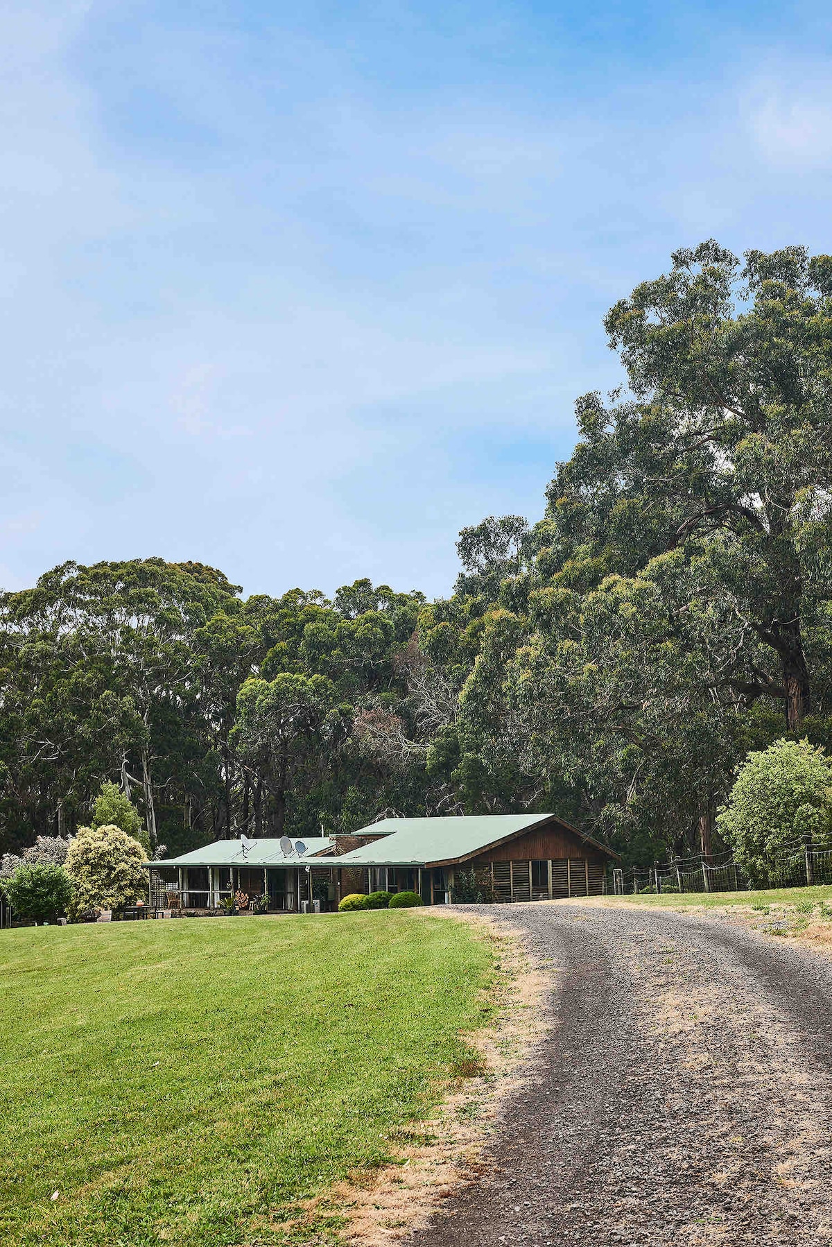 A gravel pathway leads through lush greenery to a spacious retreat nestled among tall trees. The house features a sloped roof and a wide porch, surrounded by manicured lawns and expertly tended gardens, creating a serene environment.