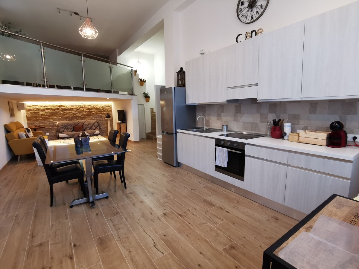 An open kitchen and dining area features light wood floors and modern cabinetry. A dining table is set for six, while a cozy seating area is visible in the background. Natural light fills the space, highlighting the contemporary design and neutral color palette.