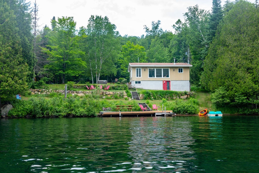 A charming chalet is situated by a clear lake, with a spacious green lawn featuring several lounge chairs. A wooden dock extends into the water, alongside canoes and kayaks, creating an inviting space for outdoor activities.