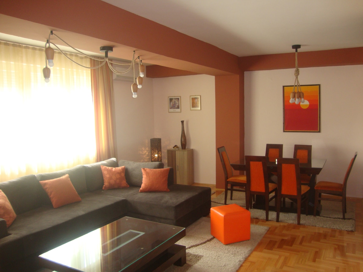 The living room features a comfortable sectional sofa adorned with orange cushions. A glass coffee table sits on a soft rug, while a dining area is visible with a wooden table and orange-accented chairs. Natural light floods the space through large windows.