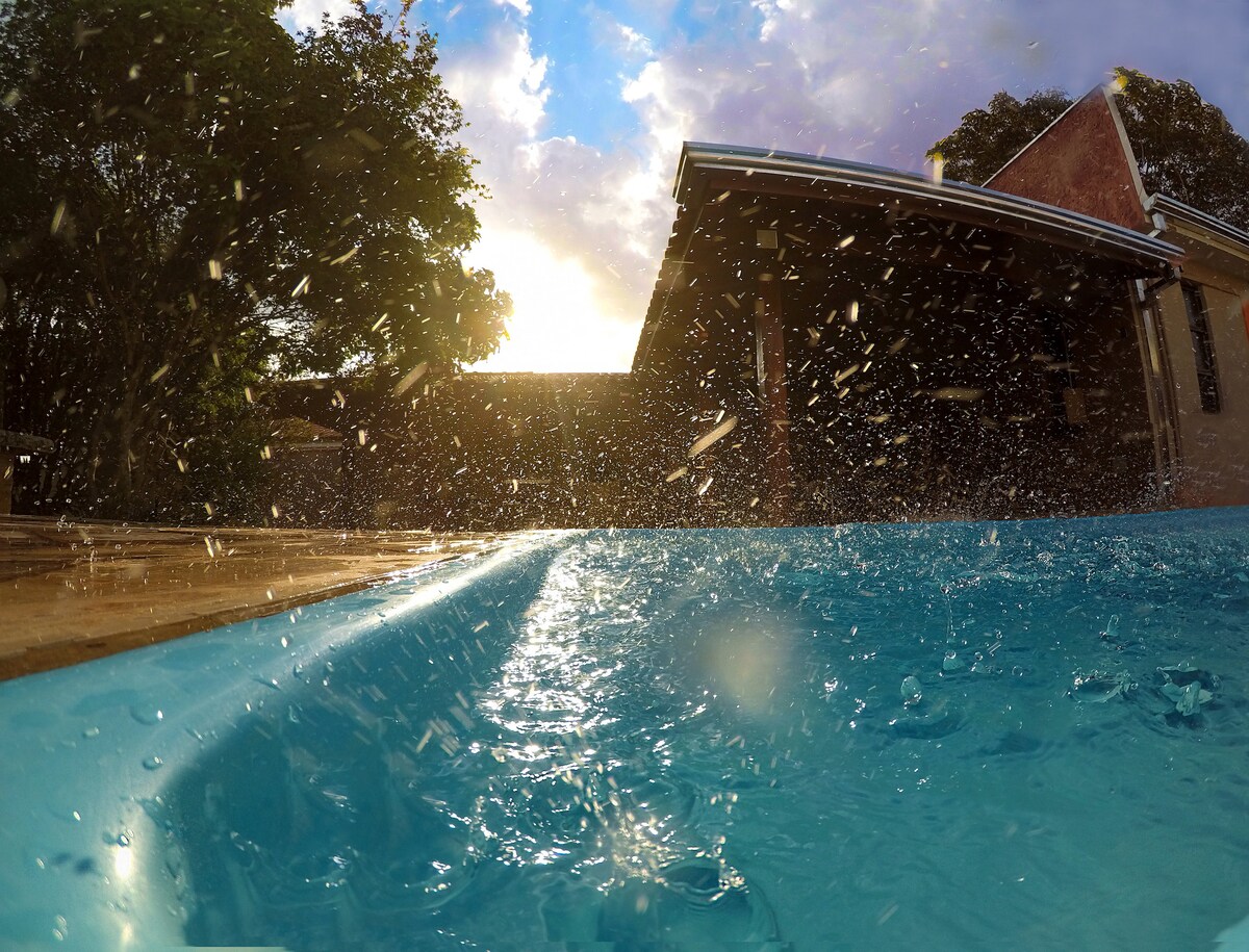 The image captures a refreshing swimming pool with water splashing against the edge. Sunlight filters through trees in the background, creating a serene ambiance. A portion of the structure is visible, indicating the integration of indoor and outdoor spaces.