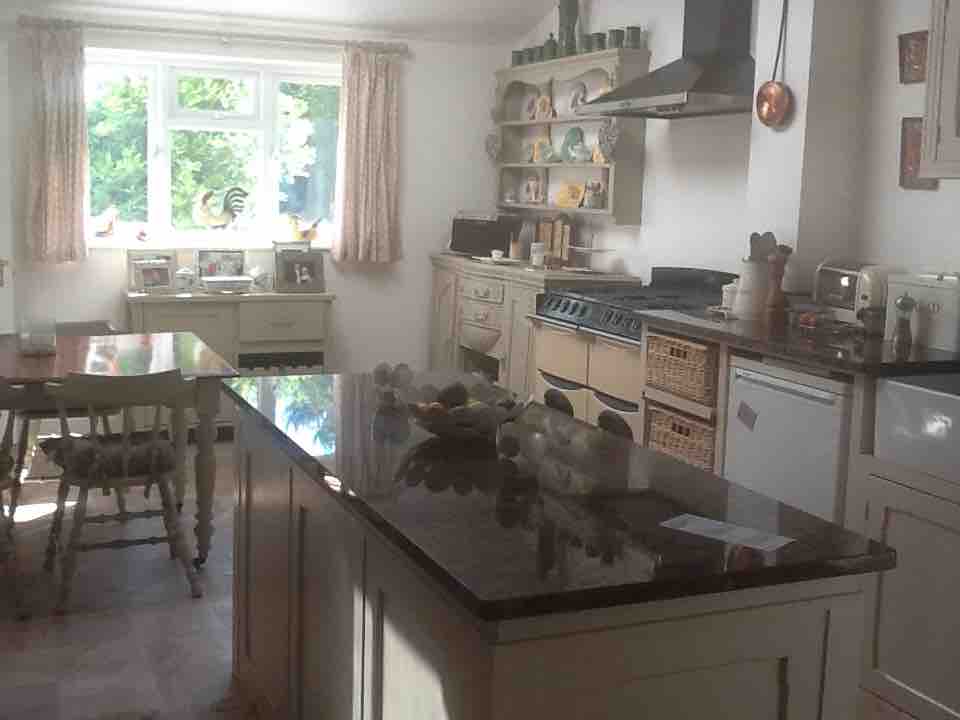 A large kitchen area is depicted, featuring a central island with a dark countertop. Cabinets display various dinnerware, while a gas stove and refrigerator are visible. Natural light streams through a large window adorned with light curtains, complementing a cozy dining table and chairs.