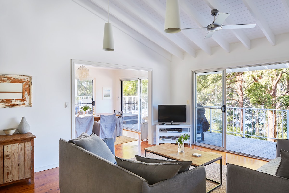 An open-plan living area is presented with high, white beamed ceilings, featuring a cozy seating arrangement and a coffee table. Large glass sliding doors offer views of the deck and surrounding trees, while a ceiling fan enhances airflow. A television is positioned on a stand nearby.