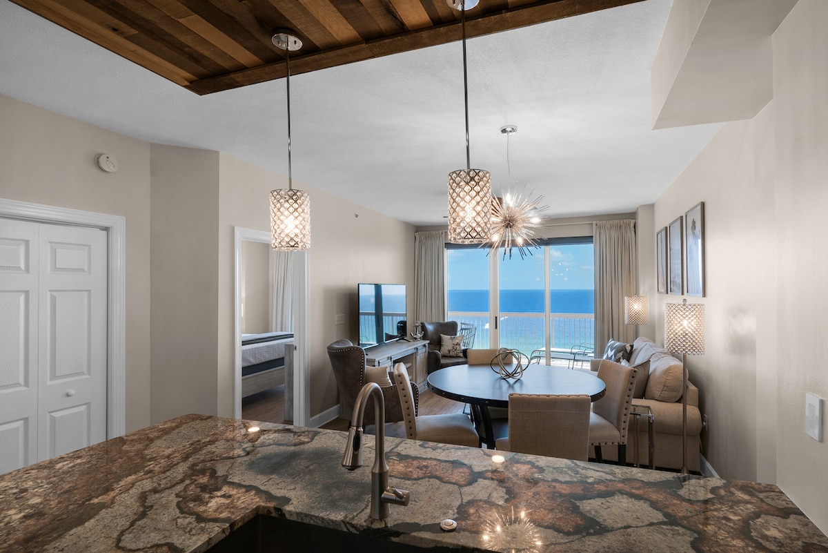 A modern kitchen with granite countertops is showcased, featuring pendant lighting above the island. A dining table sits in the foreground, accompanied by plush chairs. Through large windows, panoramic ocean views can be seen, enhancing the bright, airy ambiance of the living space.