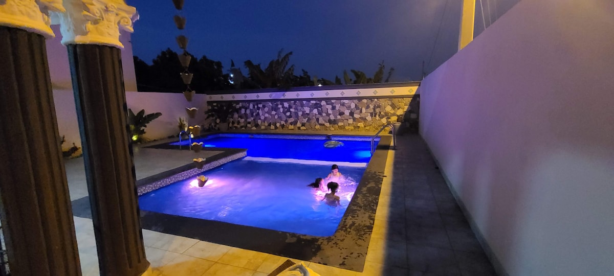 A private pool is illuminated at night, showcasing vibrant blue water. Surrounding the pool are decorative stone walls and lush greenery, creating a tranquil environment. Soft lighting enhances the serene atmosphere. The pool area is spacious, accommodating several guests comfortably.