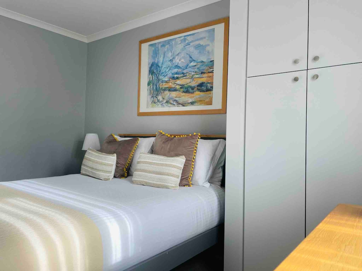 A serene bedroom features a spacious bed adorned with soft pillows. Light grey walls are complemented by a large piece of artwork above the bed. Minimalistic cabinetry offers ample storage, while a bedside lamp adds a subtle touch of illumination.