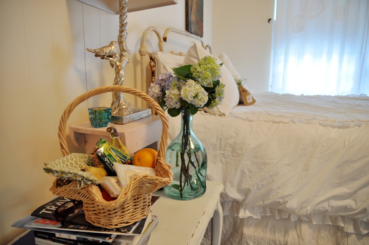 A cozy sleeping area is furnished with a white bed, complemented by a delicate lamp and decorative artwork. A woven basket holds snacks and fruits, while a vase showcases fresh flowers. Magazines are casually stacked on a nearby table, adding to the relaxed ambiance.