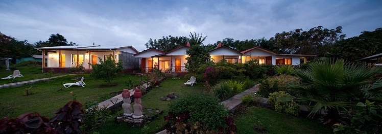 A spacious garden surrounds a single-story accommodation featuring multiple entrances adorned with warm lighting. Lush greenery and colorful plants reflect the natural beauty of the area. Comfortable lounge chairs are positioned on the grass, inviting relaxation amidst the tranquil setting.