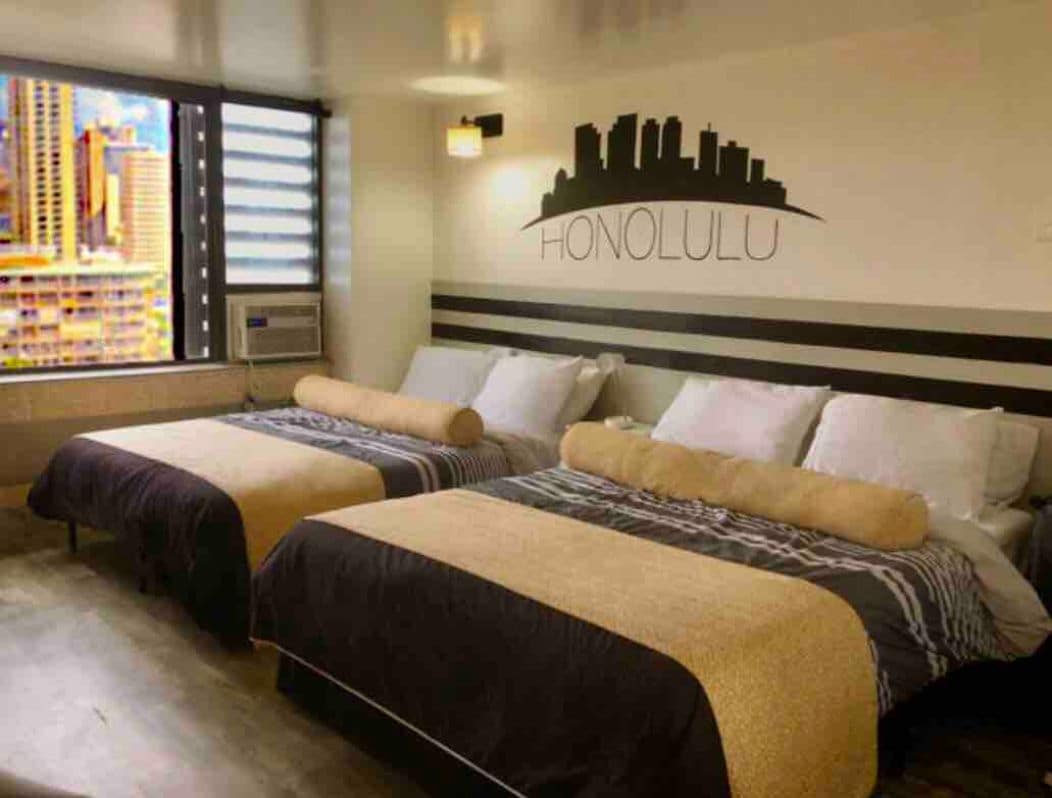 The room features two spacious queen beds adorned with neutral-toned bedding. A city skyline mural is displayed above the beds, complemented by large windows allowing natural light and offering a view of the urban surroundings.