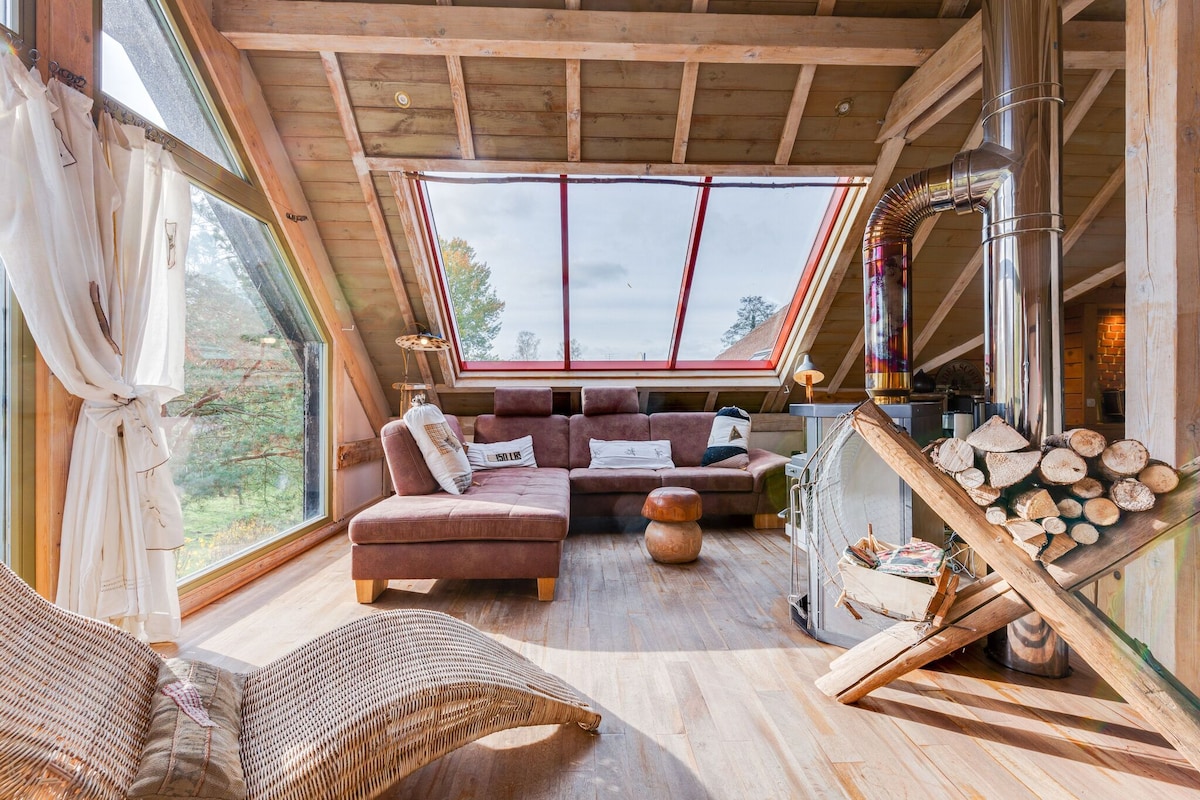 A spacious living area is illuminated by large angled windows, offering views of the outdoor scenery. A brown sectional couch faces a cozy fireplace, while a small round table and a decorative lounge chair add warmth to the space. Stacked firewood is neatly arranged nearby.
