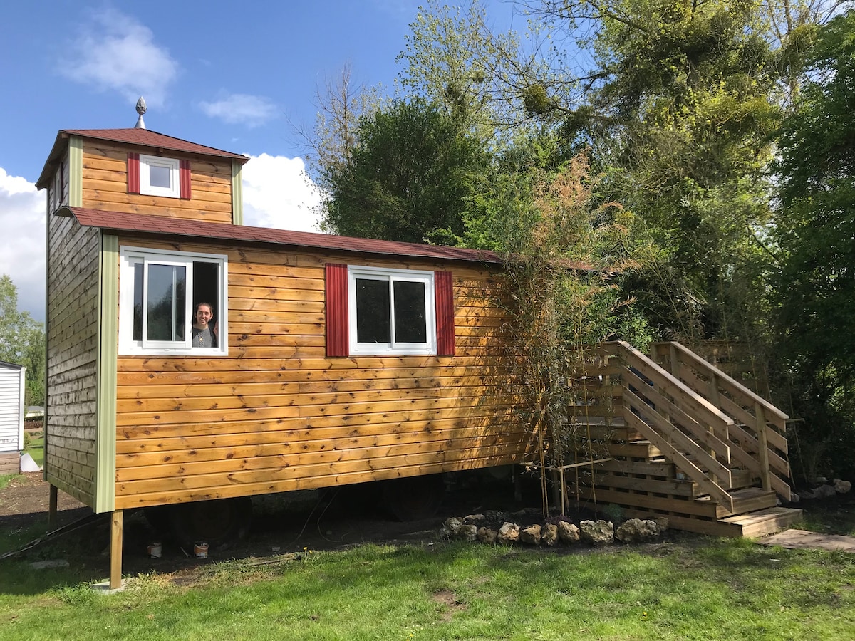 A rustic mobile cabin with a wooden exterior is set against a backdrop of greenery. A staircase leads to the entrance, while a small tower features a window with red shutters. Bright blue skies and scattered clouds create a relaxed atmosphere.