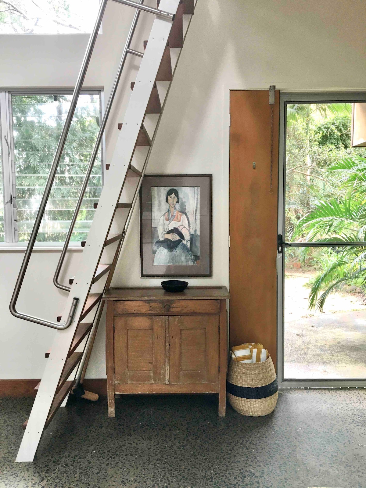 A staircase leads to the mezzanine bedroom, positioned beside a framed artwork featuring a portrait. A wooden cabinet is placed below the stairs, complemented by a black bowl atop it. Natural light pours in through large windows, illuminating the entrance area.