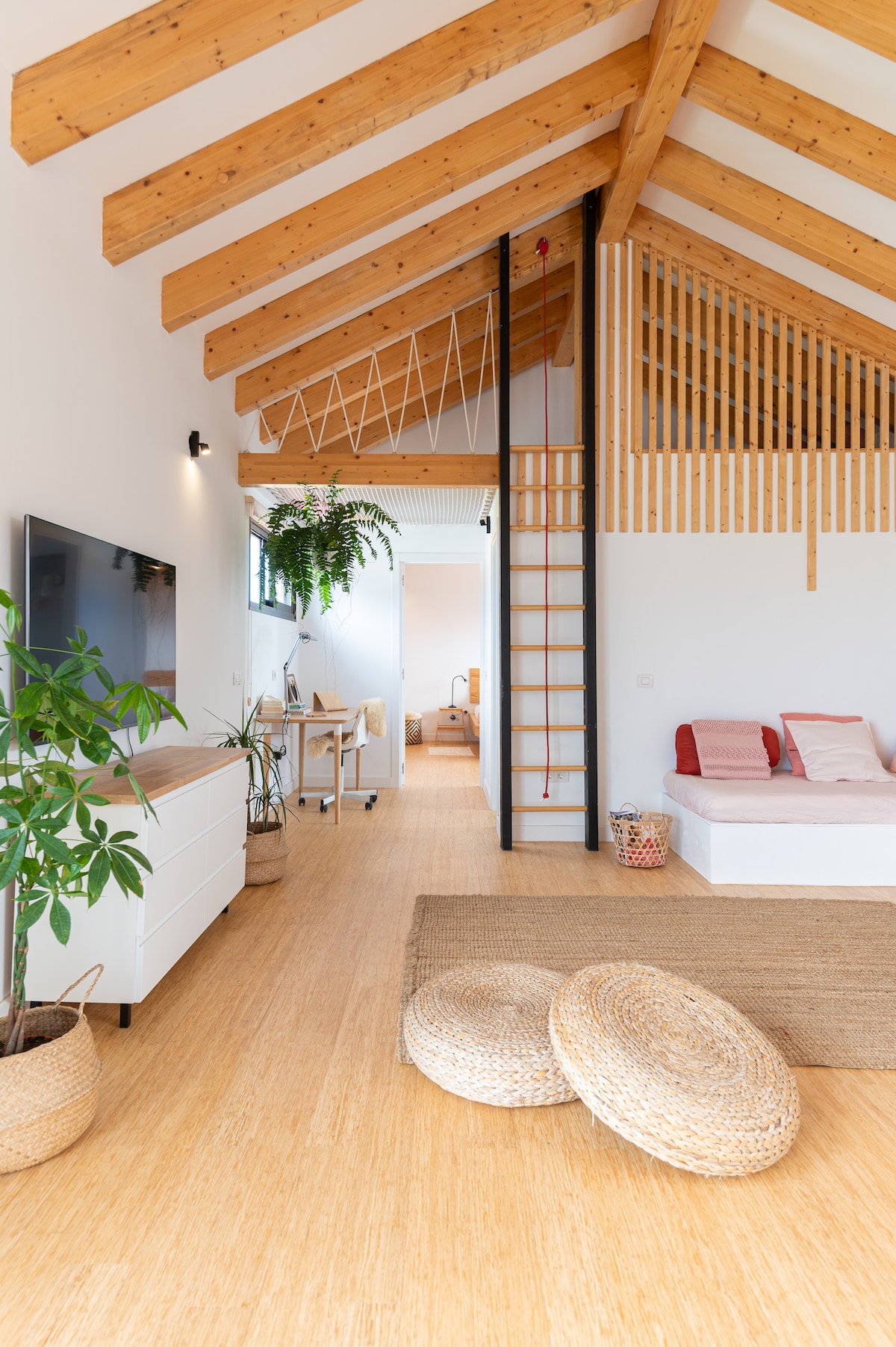 An open living area features high, wooden beams and bamboo flooring. A white dresser is placed against one wall, while a television is mounted nearby. A cozy seating nook is visible, along with a ladder leading to an upper loft area. Decorative plants and woven baskets enhance the natural decor.