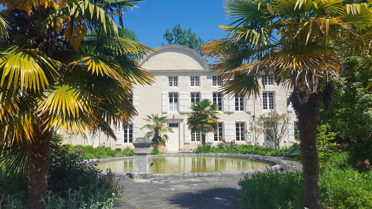 The historic château is framed by lush palm trees and vibrant foliage, creating a serene ambiance. A tranquil pond reflects the building’s classic façade, while large windows invite natural light into the interior. The scene captures the charm of the surrounding gardens.