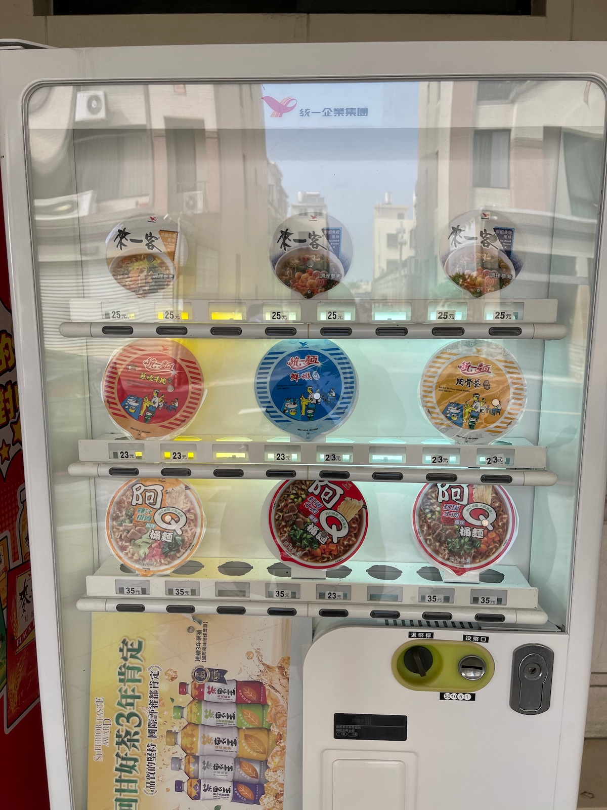 A vending machine displays a variety of packaged food items, including instant noodles and snacks. Clear labels indicate the prices on each shelf, with assorted flavors visible. The machine is mounted against a street backdrop, showcasing a quiet urban environment.