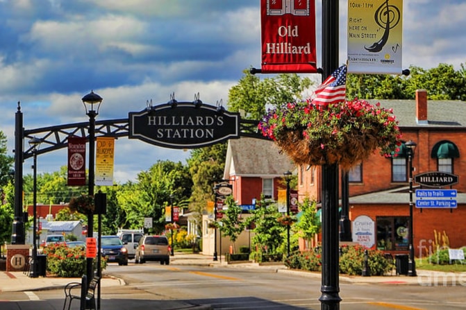 The image captures a scenic view of Hilliard's Station, featuring decorative street signs and a hanging floral arrangement. The historic charm of Old Hilliard is highlighted by the surrounding buildings and trees, while an American flag waves from a lamppost, contributing to the neighborhood's welcoming atmosphere.