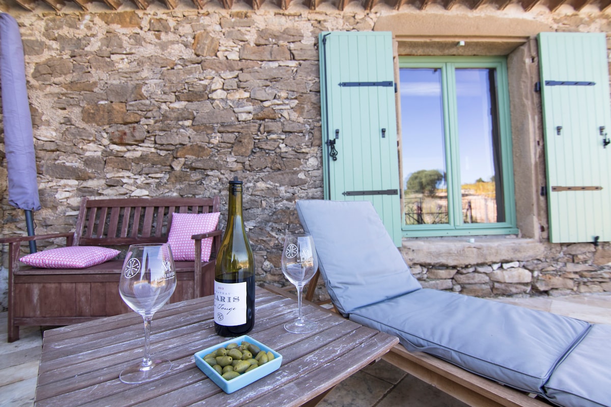 An outdoor terrace is framed by rustic stone walls, featuring a small wooden table set with a bottle of wine and two glasses. A comfortable lounge chair and a sofa with soft cushions are included, with green shutters visible on the window in the background.
