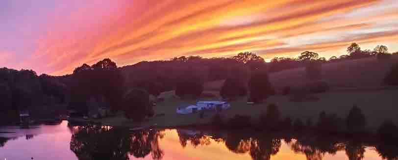 A stunning sunset casts vibrant hues across the sky, reflected on the calm lake's surface. Lush greenery surrounds the water, with a glimpse of a white residence in the background, complementing the natural beauty of the serene landscape.