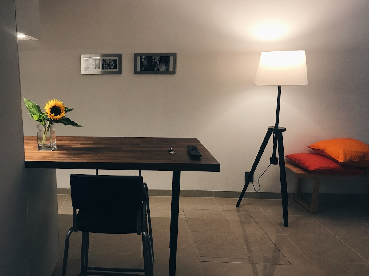 A simple workspace is presented with a wooden table and a metal chair. A small vase with a sunflower adds a natural element. A floor lamp provides warm lighting, while a sitting area features a stool and colorful cushions.