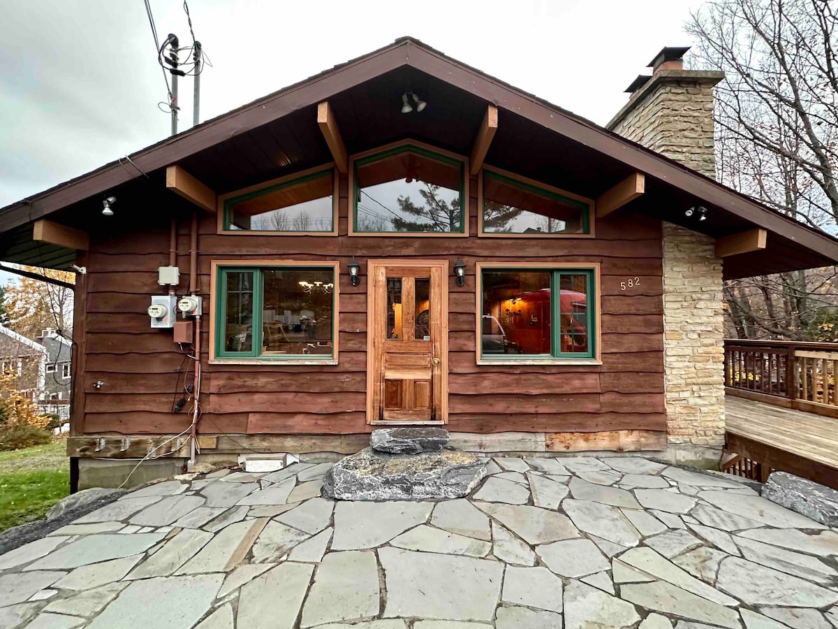 The cabin's exterior features a blend of wood and stone elements, with large windows that provide ample natural light. A wide stone path leads to the entrance, while a wooden deck extends from the front door, surrounded by grassy areas.