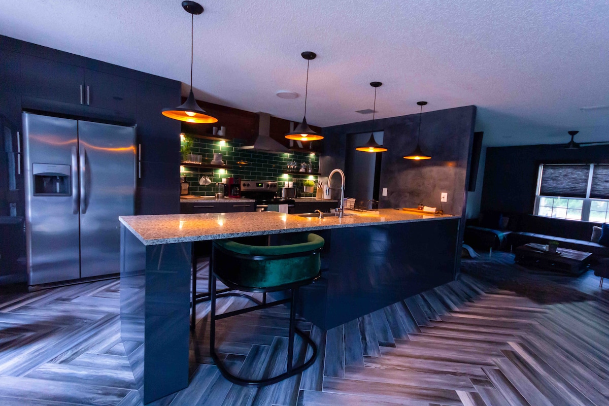 The kitchen features a large island with a granite countertop, accompanied by modern bar stools. Pendant lights illuminate the space, where green tiles add a touch of color. Stainless steel appliances and stylish cabinetry are visible, enhancing the functional design of this fully equipped kitchen.