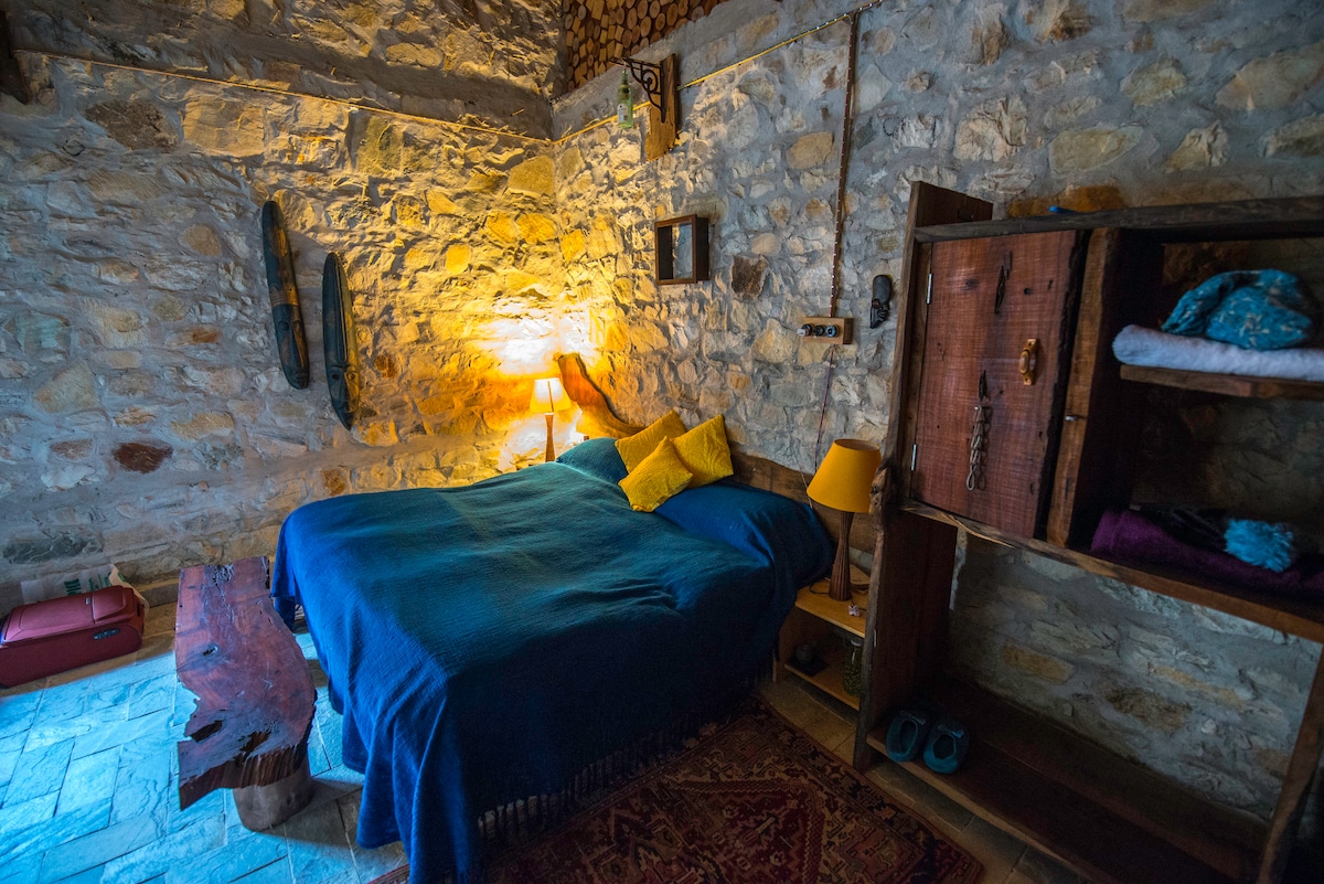 A warm bedroom is highlighted by a spacious bed draped in a blue blanket. Stone walls create a rustic ambiance. Beside the bed, bedside tables with lamps provide soft lighting. A wooden bench serves as a seating area, and shelves offer storage with folded linens.