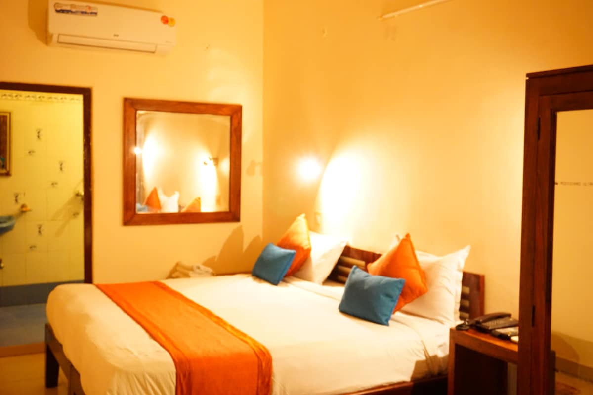 A bright and inviting room features a spacious bed adorned with orange and blue cushions. Soft lighting is provided by wall sconces and a mirror reflects the warm hues. A wooden wardrobe and a small desk enhance the functional layout, while a hint of a modern bathroom is visible in the background.