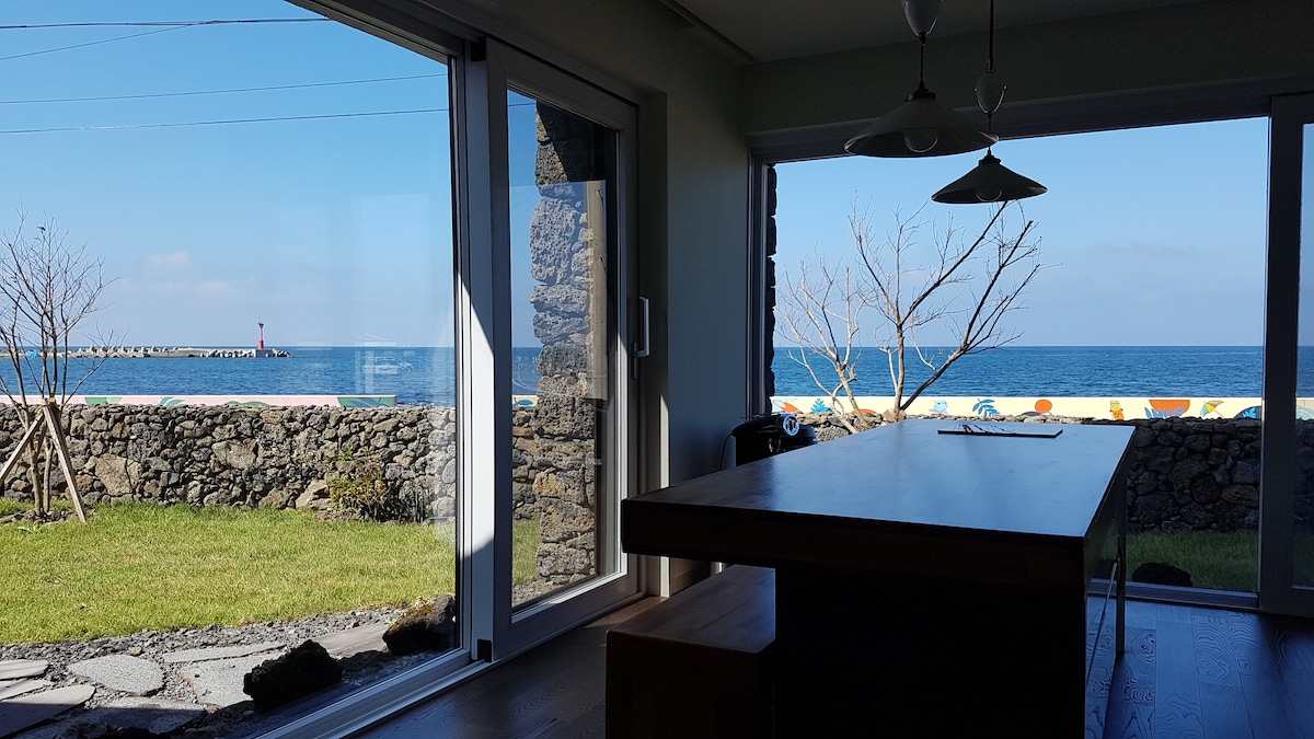 A spacious dining area is shown with large glass doors that open to stunning ocean views. The scene features a wood dining table, minimalist decor, and a glimpse of the lush outdoor space, framed by a stone wall and a clear blue sky.