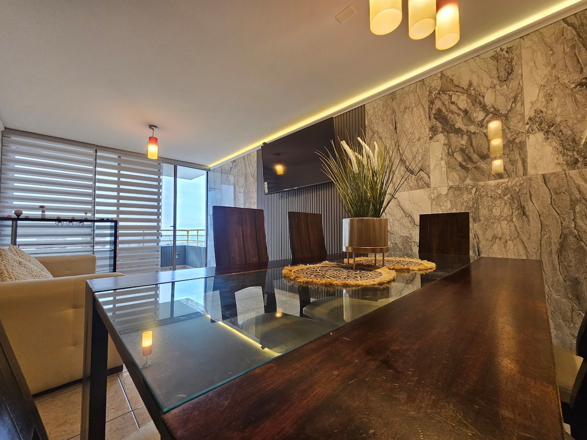 A dining area features a sleek glass table surrounded by wooden chairs. A modern centerpiece with greenery rests on the table. Light from overhead fixtures provides illumination, while large windows offer a view of the outside.
