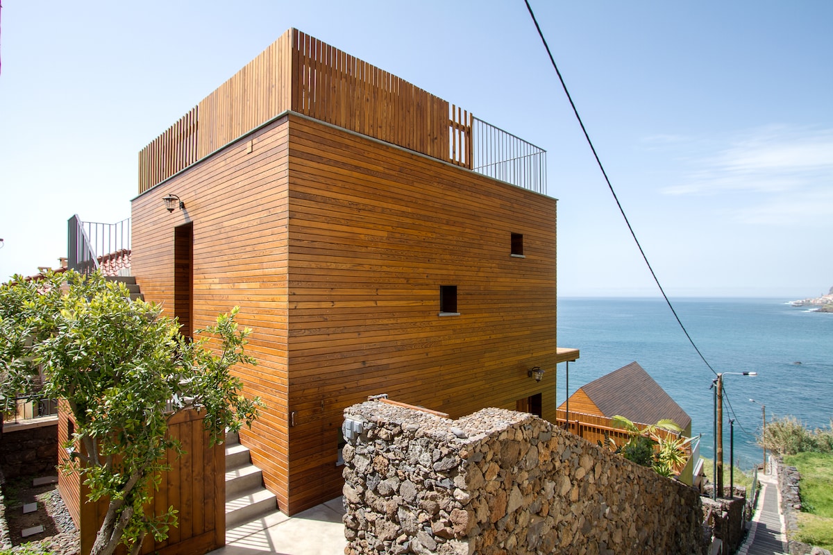 A modern wooden structure is featured, showcasing a private terrace that offers views of the ocean. The natural wood siding complements the serene surroundings, while a stone pathway leads up to the entrance. Clear skies enhance the tranquil coastal atmosphere.