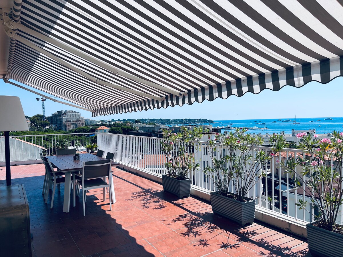 A spacious terrace is shown, featuring a dining table with chairs underneath a striped awning. Planters with greenery line the edges, offering a view of the blue Mediterranean waters and distant boats. The setting reflects a serene outdoor space for relaxation and dining.
