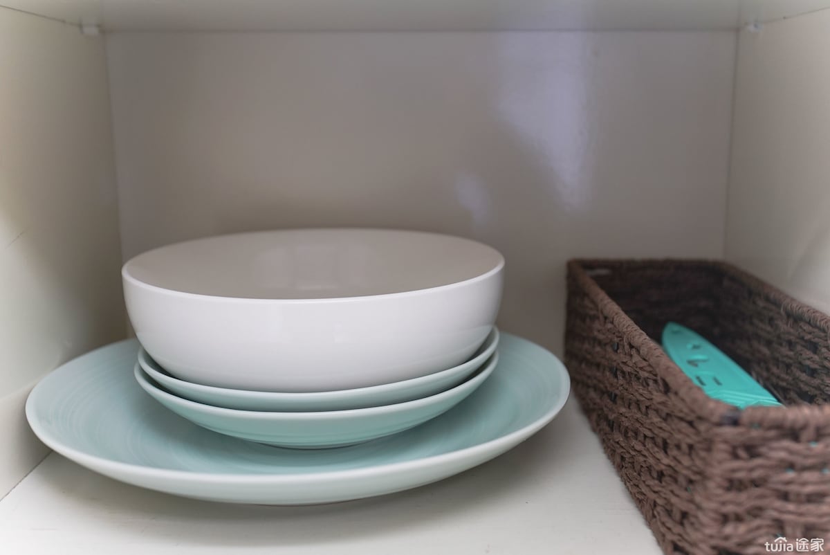 A neatly organized cabinet displays a stack of flat plates in soft green tones and a large white bowl. A woven basket containing utensils is also visible, enhancing the kitchen storage space.