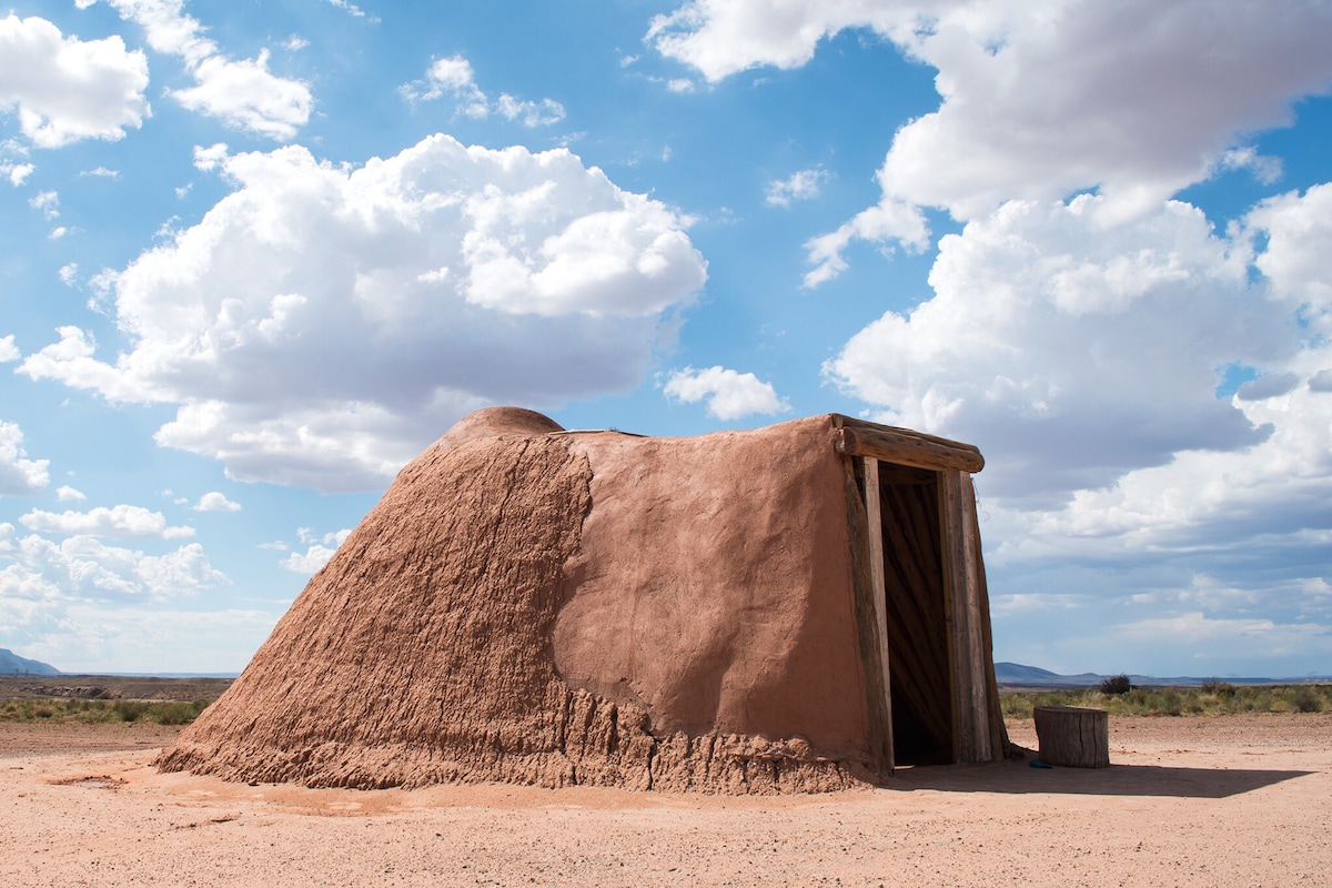 A traditional earthen hogan is shown, characterized by its rounded shape and wooden door. The exterior features natural earth tones, blending with the surrounding desert landscape under a bright blue sky dotted with fluffy white clouds.