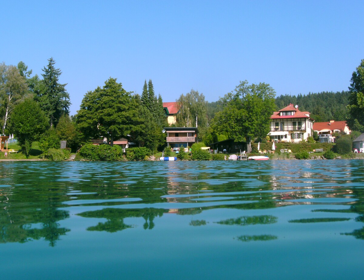 A view from the water's edge showcases a serene lakeside scene, featuring well-maintained gardens and charming homes set against a backdrop of lush trees. The clear blue sky reflects on the gentle waves of the lake.