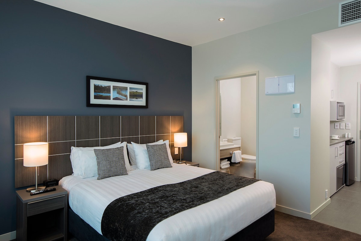 A modern studio apartment features a spacious bed with neutral bedding and decorative pillows. Warm lighting is provided by bedside lamps. A doorway leads to a bathroom, while a kitchenette area is visible in the background, reflecting a contemporary design.