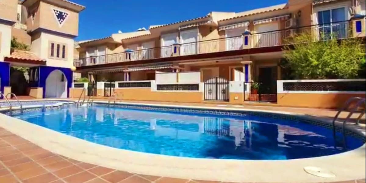 A community swimming pool is showcased with clear blue water reflecting the surrounding architecture. The area is framed by tiled deck space, leading to several residential units above. Lush greenery is present near the side, enhancing the outdoor environment.
