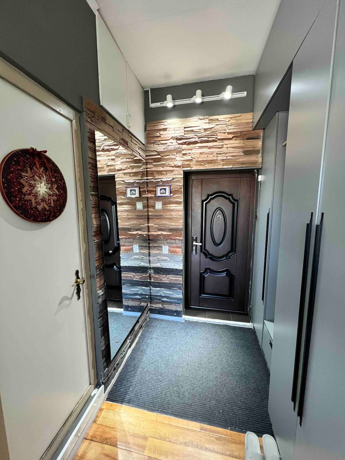 A welcoming entrance area features a dark wooden door and a full-length mirror on one side. The walls showcase textured stone tiles, complemented by soft lighting. A dark rug lines the floor, leading towards the door, enhancing the sense of space and openness.