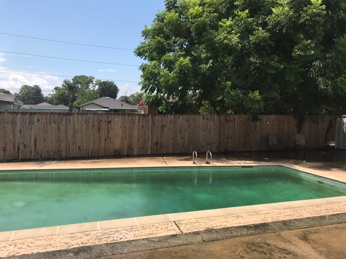 The pool area features a twelve-foot deep pool framed by a concrete deck, partially shaded by a lush tree. A wooden fence provides privacy, while two chairs sit beside the pool, ready for relaxation. The clear water invites swimming during warmer months.