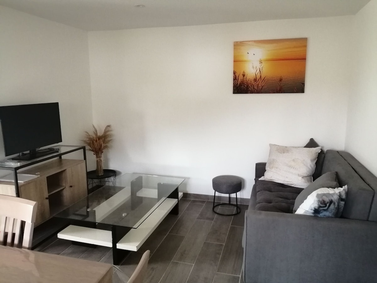 A living area is presented, featuring a comfortable gray sofa and a clear glass coffee table. A television is positioned atop a light wood console. A small dark stool sits nearby, and a piece of wall art depicting a sunset adds a touch of color.