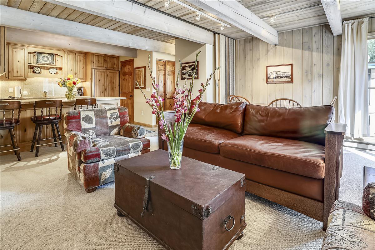 A cozy living space features a brown leather sofa and an upholstered armchair with a patterned design. A wooden coffee table sits at the center, adorned with a floral arrangement. Soft lighting enhances the warm wood accents on the walls and ceiling.