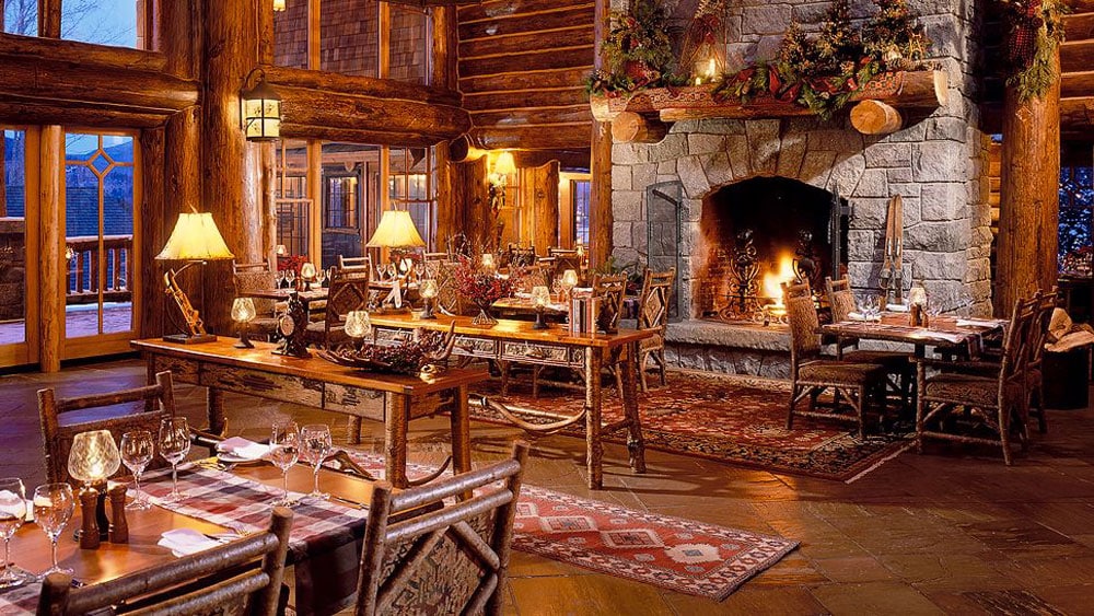A rustic dining area showcases handcrafted wooden tables surrounded by log-style chairs. A large stone fireplace serves as a focal point, adorned with seasonal decor. Soft lighting from table lamps and overhead fixtures creates a warm ambiance, complemented by decorative rugs underfoot.